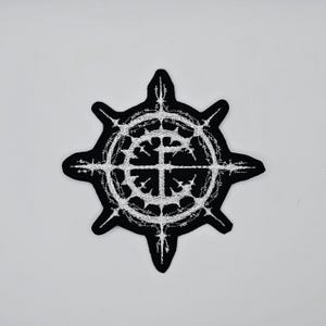 May include: Black and white embroidered patch featuring a stylized eight-pointed star design. The central element is a detailed circular pattern with a cross-like structure. The patch has a black border and is likely for clothing or accessories.