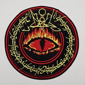 Könnte beinhalten: Gestickter Aufnäher mit rotem Auge und Flammen in der Mitte, umgeben von Schwarz mit goldfarbenen Buchstaben. Das Design umfasst ein Pentagramm und weitere Symbole, vor weißem Hintergrund.