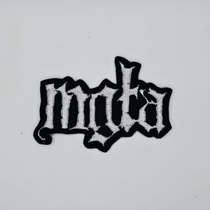 Pode incluir: Patch bordado em preto e branco com a palavra "mnta" em fonte gótica. O patch tem uma borda preta e provavelmente foi projetado para ser costurado ou passado a ferro no tecido. A escrita é branca.