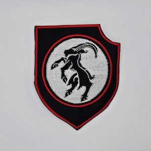 Puede incluir: Parche bordado en negro y rojo con la silueta negra de un macho montés dentro de un círculo blanco. El parche tiene forma de escudo con un borde rojo.