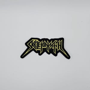 Puede incluir: Parche bordado en negro y dorado con el nombre de la banda "SKELETONWITCH" en una fuente angular estilizada. El parche tiene un fondo negro con hilo dorado para las letras y el borde.