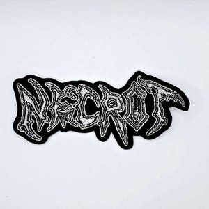 以下が含まれることがあります： 白で刺繍された「NECROT」の文字が入った黒いパッチ。ゴシック様式のフォントを使用。パッチはユニークで不規則な形状をしており、テキストは質感のある、使い古された外観で、ジャケットやバッグに適しています。