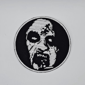 May include: A circular black and white patch featuring a zombie face design. The zombie face is white with detailed shading, set against a black background. The patch is likely for clothing or accessories.