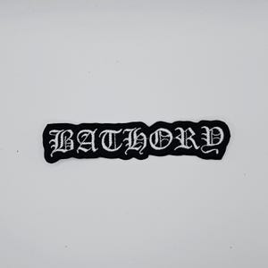 May include: Black embroidered patch with the word "BATHORY" in a gothic font. The patch has a black border and white lettering. The patch is rectangular in shape and is likely designed to be sewn or ironed onto clothing or accessories.