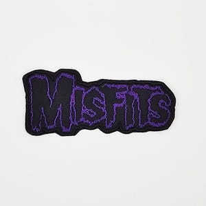 Op de afbeelding: Een zwart embleem met het woord "MISFITS" geborduurd in paars, met een gekarteld, omlijnd lettertype. Het embleem heeft een zwarte rand en is geplaatst tegen een witte achtergrond. Ideaal voor jassen, tassen of andere kleding.