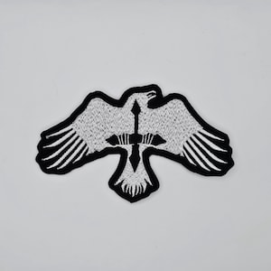 May include: Embroidered patch depicting a white bird with outstretched wings, a black border, and a black cross design. Suitable for application on clothing or accessories. The patch is in a bird shape.