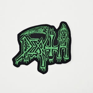 Op de afbeelding: Een zwart embleem met het woord "DEATH" in een groene, gotische letter. Het embleem heeft een onregelmatige vorm en is waarschijnlijk bedoeld voor kleding of accessoires. Het ontwerp bevat een kruis en andere gestileerde elementen.