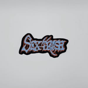 Pode incluir: Patch bordado com o texto "Sick Beast" em azul claro, contornado em laranja, sobre um fundo preto. O patch tem uma forma irregular e provavelmente foi projetado para ser passado a ferro ou costurado em roupas ou acessórios.