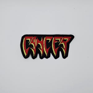 Puede incluir: Parche bordado con la palabra "CANCER" en letras rojas y amarillas sobre un fondo negro. El parche tiene un diseño irregular y goteante, probablemente para ropa o accesorios.