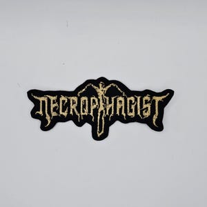 以下が含まれることがあります： ゴシック体で「NECROPHAGIST」と金色の刺繍が施された黒いパッチ。パッチにはテキストの上に様式化されたデザインがあります。このパッチは、ジャケットやバッグ用です。