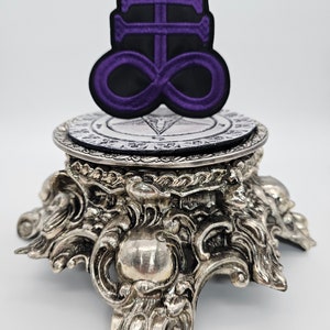 May include: Purple and black embroidered patch of the Leviathan symbol, a cross with an infinity symbol, on a silver decorative stand.