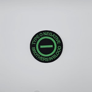 以下が含まれることがあります： 緑色の刺繍のテキストとデザインが施された、円形の黒いパッチ。テキストは上部に「TYPE O NEGATIVE」、下部に「BROTHERS IN BLOOD」と書かれています。緑色の円が黒い水平バーを囲んでいます。