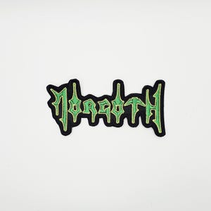 Puede incluir: Parche bordado con la palabra "FORGOTH" en letras góticas verdes. El parche tiene un borde negro y un fondo blanco. Adecuado para chaquetas, bolsos u otras prendas.