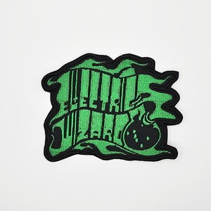 Könnte beinhalten: Ein grün-schwarzer, gestickter Aufnäher mit den Worten "Electric Wizard" in einer stilisierten Schriftart. Der Aufnäher hat einen schwarzen Rand und ein Design einer blubbernden Flüssigkeit. Der Aufnäher befindet sich auf weißem Hintergrund.