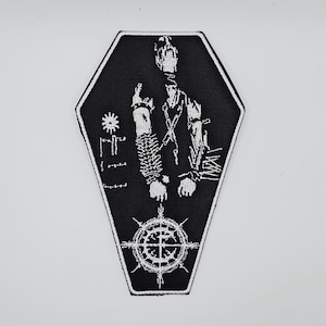 Peut inclure: Écusson brodé noir et blanc en forme de cercueil. L'écusson représente une figure squelettique avec une couronne, des chaînes et un motif circulaire en bas. Le mot "EII" est sur le côté gauche.