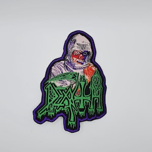 May include: Embroidered patch featuring a figure with red and purple facial details, wearing a white hood. The patch has a purple border and the word "DEATH" in green, dripping letters. The patch is on a white background.