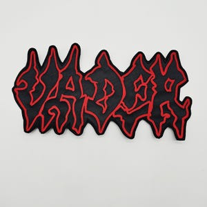 May include: A black embroidered patch featuring the word VADER in a stylised, jagged, heavy metal-inspired font. The lettering is outlined in a bold, vibrant red thread, creating a sharp contrast against the black fabric background of the patch.