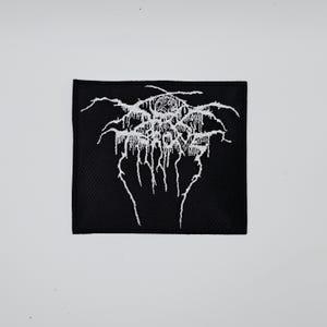Puede incluir: Parche cuadrado negro con diseño abstracto bordado en blanco. El diseño presenta un patrón estilizado complejo con un grupo central y elementos lineales en cascada. El parche es probablemente para ropa o accesorios.