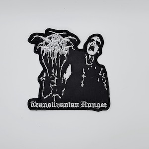 May include: A black and white embroidered patch with a screaming figure and abstract shapes. The text "Transilvanian Hunger" is displayed in a gothic font at the bottom of the patch. Ideal for jackets or bags.