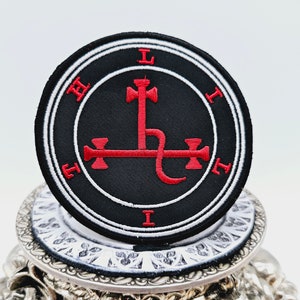 以下が含まれることがあります： 赤色の紋章が描かれた黒と白の刺繍パッチ。紋章は蛇が巻き付いた十字架。パッチは白の円形に囲まれ、白の文字で「L I T I T I」と記されている。