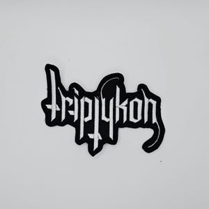 Pode incluir: Um patch bordado em preto e branco com a palavra "Triptykon" em uma fonte gótica. O patch tem uma borda preta e está sobre um fundo branco. O design é arrojado e marcante.