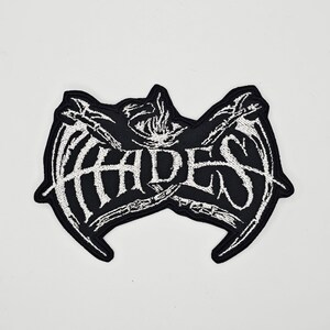 Op de afbeelding: Zwart-wit geborduurde patch met het woord "HADES" in een gestileerd lettertype. De patch heeft een ontwerp met skeletvleugels en een vlamachtig element boven de tekst. Geschikt voor kleding of accessoires.