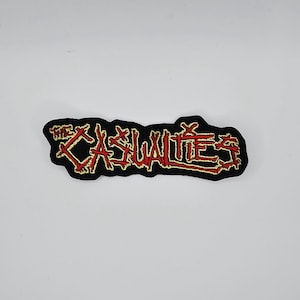 May include: Black embroidered patch featuring the words "THE CASUALTIES" in red and gold thread. The patch has an irregular shape and is designed to be sewn onto clothing or accessories.