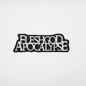 Può includere: Una toppa ricamata in bianco e nero con la scritta "FLESHGOD APOCALYPSE" in un font stilizzato. La toppa ha un bordo nero ed è posta su uno sfondo bianco.