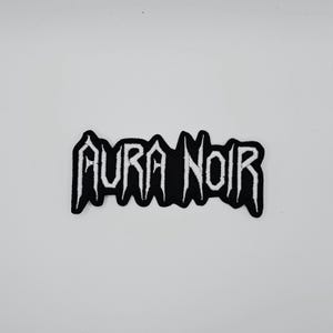 May include: Black and white embroidered patch with the text "AURA NOIR" in a gothic font. The patch has a black background and white stitching, with a jagged border. The patch is likely for clothing or accessories.