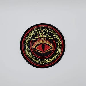 May include: Circular black patch with a red trim, displaying a red eye design with flames and gold lettering. The eye is central, encircled by gold text. This patch is likely for application on clothing or accessories.