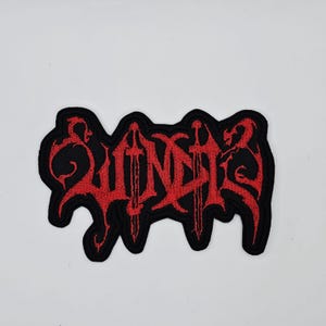 Może przedstawiać: Czarna, haftowana naszywka z napisem "SUNDER" w czerwonym, stylizowanym kroju pisma. Projekt zawiera trzy miecze i elementy dekoracyjne. Naszywka ma czarną obwódkę i jest przeznaczona na odzież lub akcesoria.