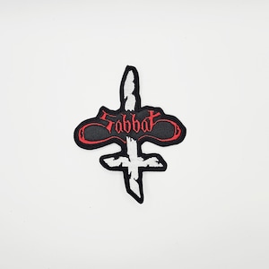 Op de afbeelding: Geborduurde patch met een gestileerd kruisontwerp en het woord "Sabbat" in rood schrift. De patch heeft een zwarte rand en een wit interieur, geschikt voor jassen, tassen of andere stoffen items.