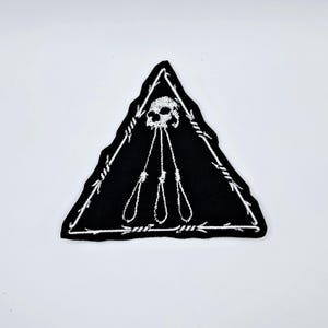 Puede incluir: Parche triangular negro con un diseño de calavera blanca y lazos corredizos. El parche está bordeado con alambre de púas blanco. La calavera está en la parte superior del triángulo, con tres lazos corredizos debajo.