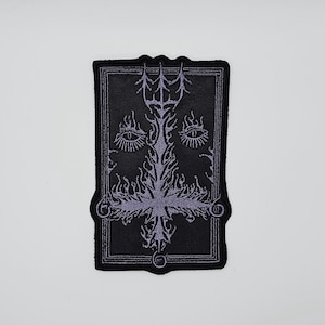 May include: A black embroidered patch featuring a symmetrical design with a central cross-like shape, two eyes, and ornate details. The design is in a lighter shade of purple, set against a black background.