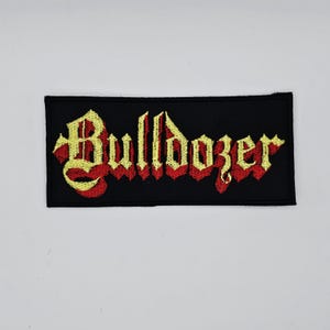 Puede incluir: Parche bordado con la palabra "Bulldozer" en fuente gótica. El texto es amarillo con sombra roja, sobre un fondo rectangular negro. Adecuado para ropa o accesorios.