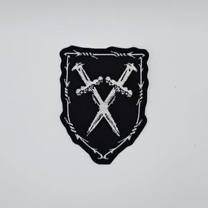 May include: Black patch featuring a white embroidered design of two crossed swords within a shield shape. The shield is outlined with a white barbed wire design. Suitable for clothing or accessories.