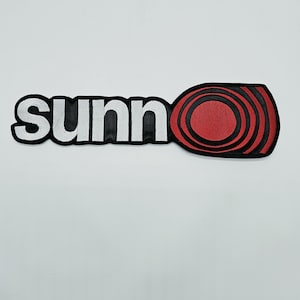 Puede incluir: Un parche bordado que presenta el texto en minúsculas blancas sunn seguido de un gráfico circular rojo y negro. El diseño está delineado con hilo negro, creando una forma que combina letras con un icono estilizado.