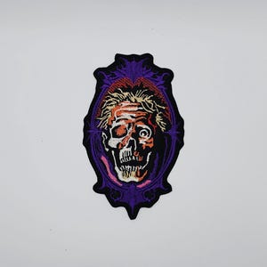 May include: Embroidered patch featuring a stylized skull with exposed brain matter and a single eye, set within a purple, ornate frame. The skull has orange, black, and white details. The patch is likely for clothing or accessories.