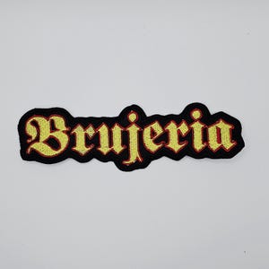 May include: Embroidered patch with the word "Brujeria" in yellow gothic lettering, outlined in red, on a black background. The patch has a black border and is likely for clothing or accessories.