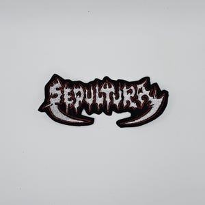 May include: Embroidered patch featuring the word "SEPULTURA" in white, stylized, gothic lettering. The patch has a black background with a red border, and a jagged, flame-like design. The patch is likely for clothing or accessories.
