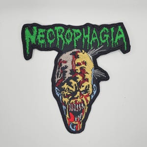 Può includere: Toppa ricamata con la scritta "NECROPHAGIA" in verde, sopra un volto di zombie. Il volto dello zombie presenta dettagli rossi, gialli e blu, con denti scoperti e un aspetto in decomposizione. La toppa ha un bordo nero.