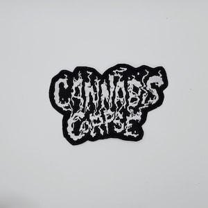 Peut inclure: Écusson brodé noir et blanc avec l'inscription "CANNABIS CORPSE" dans une police gothique stylisée. Le patch a un contour noir et un intérieur blanc, avec une forme irrégulière.