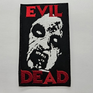 Puede incluir: Un parche de tela negro con bordado blanco de una cara de zombi y bordado rojo de las palabras "EVIL DEAD".