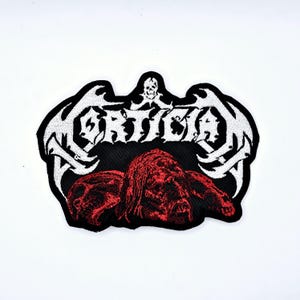 May include: Embroidered patch with the word "Mortician" in white, gothic-style lettering, above a red and black graphic. The patch has a black border and a skull at the top. The patch is likely for a jacket or bag.
