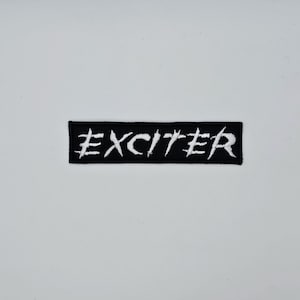 May include: A black rectangular patch featuring the word "EXCITER" in white, embroidered lettering. The patch is set against a white backdrop. The embroidery has a textured finish.