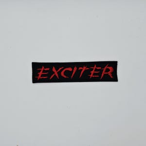 May include: A black rectangular patch featuring the word "EXCITER" in red, embroidered lettering. The patch is designed to be sewn or ironed onto fabric. The background is plain white.