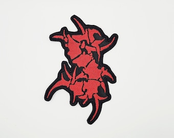 Puede incluir: Parche bordado con un diseño abstracto estilizado en rojo y negro. El parche tiene una forma compleja y dentada con un borde negro, sobre un fondo blanco. El diseño parece ser un símbolo o logotipo.