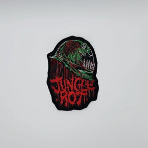 Peut inclure: Écusson brodé avec un fond noir et les mots "JUNGLE ROT" en rouge. L'écusson présente un motif vert et rouge d'un casque avec un symbole de paix et un crâne.