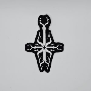 May include: A black and white embroidered patch featuring a stylized cross design. The patch has a black background with a white, intricate, tree-like pattern. The design is symmetrical and detailed, with a clean, crisp finish.