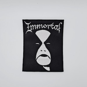 May include: Black embroidered patch featuring the word "Immortal" in white gothic lettering above a white stylized face. The patch is rectangular with a black border and a white face design.